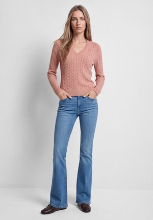 Woman wearing a pink cable knit sweater, blue flared jeans, and dark brown shoes, standing with hands in pockets against a plain background.