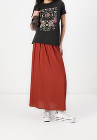 Schwarzes Grafik-T-Shirt mit floralen Design, kombiniert mit einem fließenden roten Maxikleid und weißen Turnschuhen. Tragetasche in Braun.