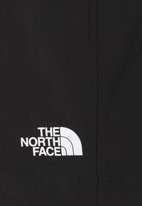 Musta kangas, missä näkyvät ompeleet, esittelee valkoista "The North Face" -logoa ja graafista puoliympyrää, mikä viittaa brändin identiteettiin.