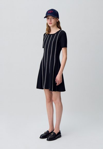 Mujer vistiendo un vestido negro con rayas blancas verticales, unos zapatos mocasines negros y una gorra de béisbol azul marino con un logotipo rojo, de pie frente a un fondo liso.