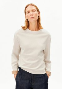 Hellgrauer Sweatshirt mit rundem Ausschnitt und langen Ärmeln, versehen mit einem umgeschlagenen Bündchen, kombiniert mit dunkelblauen Jeanshosen.