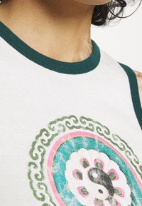 Close-up of a person wearing a white tank top with green trim and a yin-yang symbol surrounded by floral and swirl patterns on the chest.
