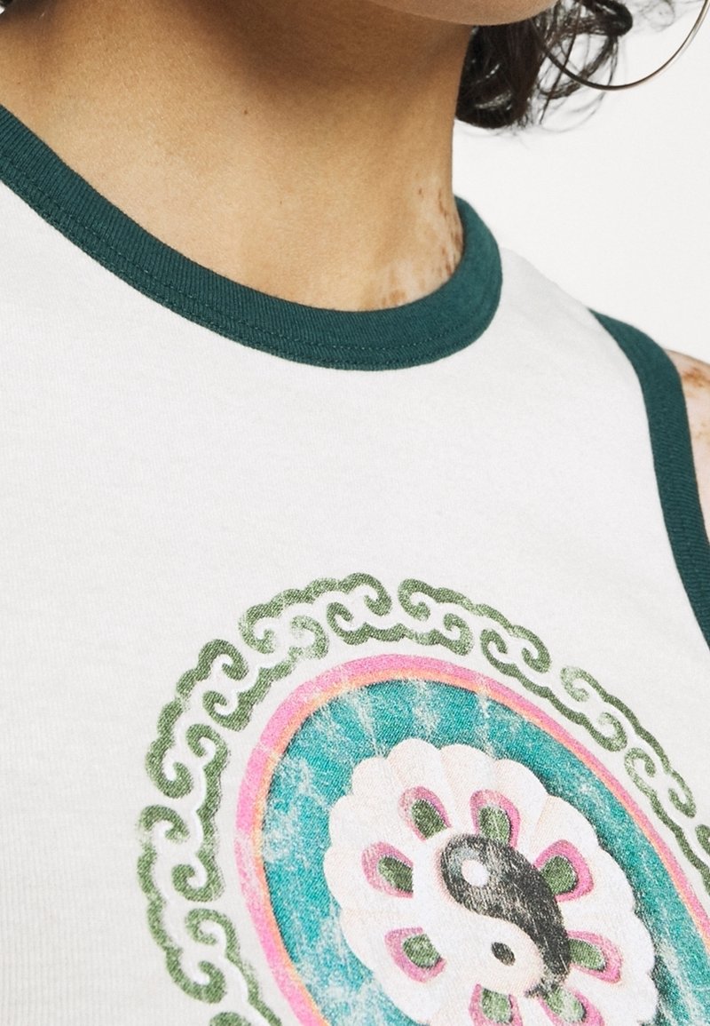 Close-up of a person wearing a white tank top with green trim and a yin-yang symbol surrounded by floral and swirl patterns on the chest.