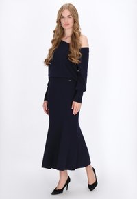 Marine blauwe off-shoulder gebreide top gecombineerd met een bijpassende lange, uitlopende rok. De outfit heeft een gladde textuur en een eenvoudig ontwerp.