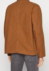 Brown cotton jacket with a straight cut, visible stitching, and a simple collar. Pairing with gray jeans, showing minimalistic design details.
