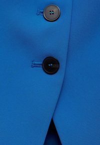 Jasnoniebieska tkanina o gładkiej teksturze, wyposażona w dwa czarne guziki przy zapięciu dla pewnego zabezpieczenia. Zauważalny kontrast między kolorami.