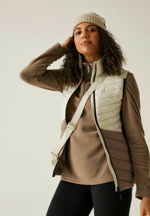 Kobieta z kręconymi włosami, ubrana w beżową dzianinową czapkę, białą i brązową pikowaną kamizelkę, brązową bluzę polarową, czarne legginsy oraz paskiem torby noszonej przez ramię.