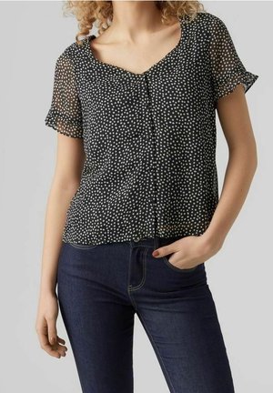 Kobieta ubrana w czarną, przezroczystą bluzkę z krótkim rękawem w białe groszki oraz ciemnoniebieskie dżinsy, z ręką w kieszeni, na jednolitym szarym tle.