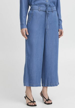 Pantalones de mezclilla anchos y azules con cintura ceñida por un cinturón, que cuentan con bolsillos laterales y una textura suave. Combinados con sandalias negras de tira.