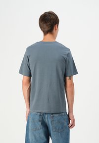 Grå t-skjorte med kort erme og rund hals, laget av mykt stoff, vist bakfra, kombinert med blå denimjeans.