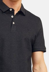 Zwarte polo gemaakt van katoen; met een knoopsluiting, korte mouwen met opgerolde manchetten, en een klein geborduurd logo op de borst.