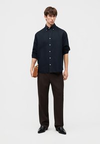 Mężczyzna ubrany w czarną koszulę na guziki, ciemnobrązowe spodnie i czarne buty, trzymający małą brązową teczkę na jednolitym tle.