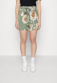 Zelené květované šortky s elastickým pasem a zavazovací detaily, zdobené velkými oranžovými a bílými květy, nošené s bílými teniskami a ponožkami.