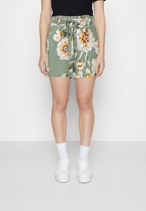 Vihreät kukkakuviolliset shortsit, joissa on joustava vyötärönauha ja solmittava yksityiskohta, suuriin oransseihin ja valkoisiin kukkiin, yhdistetty valkoisiin tennareihin ja sukkiin.