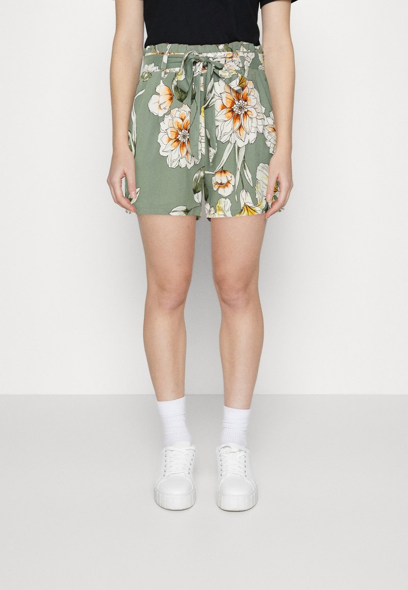 Zelené květované šortky s elastickým pasem a zavazovací detaily, zdobené velkými oranžovými a bílými květy, nošené s bílými teniskami a ponožkami.