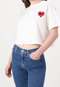 Camiseta blanca corta de algodón, con un diseño de corazón rojo en el lado izquierdo del pecho. Combinada con pantalones vaqueros de tiro alto azules.