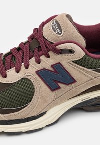 Beige Suede-Sneaker mit grünen Mesh-Akzenten und dunkelbraunen Details. Verfügt über weinrote Schnürsenkel und ein blaues Logo. Strukturierte Gummisohle.