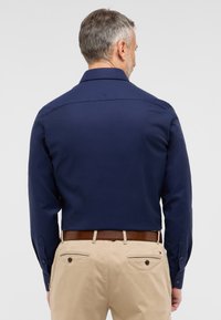 Mørkeblå langærmet skjorte med krave, der kan knappes. Bæret med lyse beige bukser og et brunt bælte, med rygbærestykke og knapdetaljer.