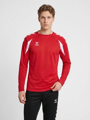Ung mand med krøllet hår iført en rød sportsbluse med lange ærmer med hvide detaljer og sorte sportbukser mod en ensfarvet baggrund.