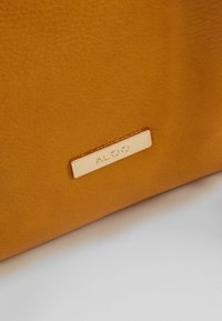 Żółta skórzana torebka z metalową plakietką w kolorze złotym z tłoczonym napisem "ALDO". Gładka faktura z delikatnym detalem ziarnistości.