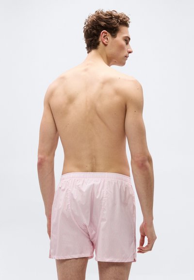 Νεαρός άνδρας με σγουρά μαλλιά που φοράει ροζ και λευκά ριγέ μποξεράκια, εμφανίζεται από την πίσω πλευρά σε απλό φωτεινό φόντο.