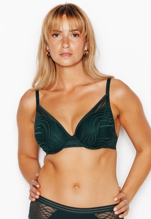 Femme blonde portant un soutien-gorge vert foncé à motifs et une culotte assortie, debout les mains sur les hanches sur fond blanc.