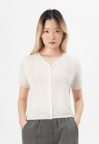 Mladá žena s dlhými ombre vlasmi má na sebe bielo button-up tričko s krátkymi rukávmi a tmavosivé nohavice, so svojimi rukami v vreckách na bielom pozadí.
