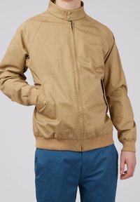 Mand iført en beige zip-up jakke med strikkede manchetter og krave, kombineret med blå bukser, stående foran en ensfarvet lys baggrund.