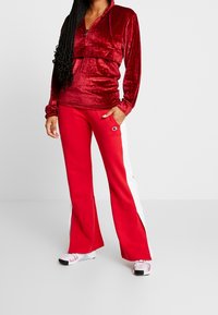 Haut zippé en velours rouge avec un design court, associé à un pantalon évasé rouge avec des bandes blanches sur les côtés et un petit logo. Finition texturée.