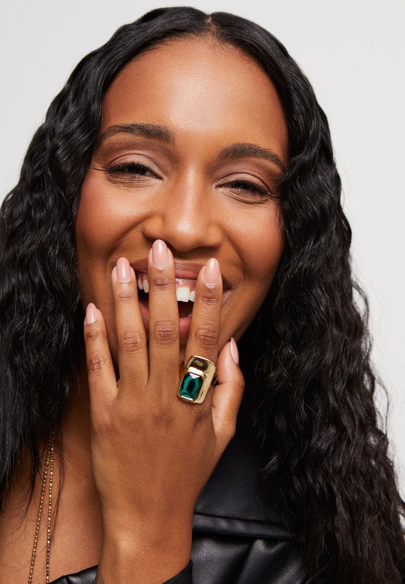 Bague en or avec une grande pierre précieuse de couleur émeraude, surface lisse et forme rectangulaire, portée sur une main avec des ongles manucurés.