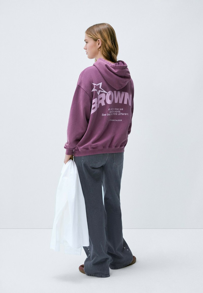 Hoodie violet avec le texte "BROWN" et un graphique d'étoile au dos. Coupe ample, porté avec un pantalon large gris foncé, tenant un objet blanc.