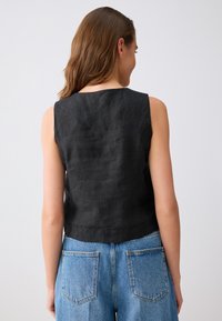 Femme vue de dos portant un haut noir sans manches et un jean en denim bleu sur un fond gris uni.