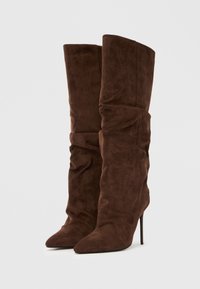 Καφέ μπότες γόνατο από σουέντ με χαλαρή σχεδίαση, μυτερές μύτες και λεπτά ψηλά τακούνια. Διαθέτουν απαλή υφή και ρυτιδωτή λεπτομέρεια.