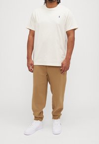 Bílý bavlněný tričko s malým modrým logem, spárované s hnědými tepláky a bílými teniskami. Pohodlný střih a casual design.