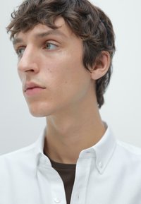 Nahaufnahme einer Person, die ein weißes Hemd mit Knöpfen trägt. Glatte Textur; kurzes, lockiges Haar; und natürliche, neutrale Hauttöne mit grünen Augen.