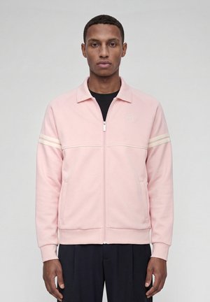 Homme portant une veste zippée rose clair avec des rayures crème sur les manches et un pantalon noir, debout devant un fond uni.