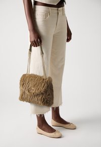 Borsa in pelliccia sintetica marrone con dettagli in metallo dorato, tenuta da una mano. Abbinata a pantaloni ampi color crema e ballerine beige.