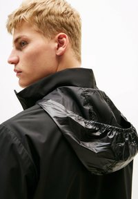 Zwarte jas met een glanzende capuchon, voorzien van een hoge kraag en elastische details. Soepele textuur en contrasterende afwerkingen.