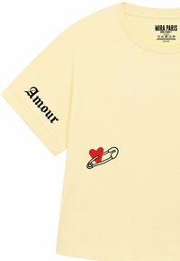 Geel katoenen T-shirt met korte mouwen. Kenmerkt zich door zwarte tekst "Amour" op de mouw en een rood hartontwerp naast een veiligheidsspeld.