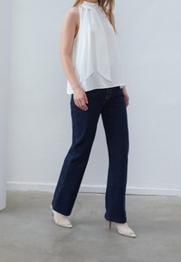 Blouse blanche sans manches avec un col montant et un devant drapé, associée à un jean évasé bleu foncé et des escarpins blancs en mesh à bout pointu.