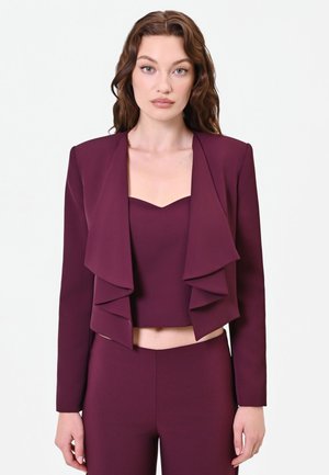 Femme aux cheveux bruns ondulés portant une veste courte violette avec des revers superposés et un pantalon taille haute assorti, debout devant un fond uni.