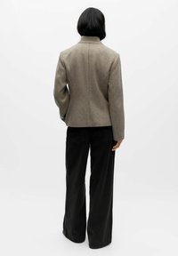 Bruine jas van een wolmix met een gestructureerde kraag, een getailleerd ontwerp en lange mouwen; gecombineerd met zwarte broek met wijde pijpen.