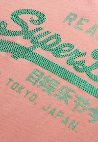 T-shirt rose avec des ornements en rhinestones verts, affichant le texte "Super" et "Tokyo, Japon" en lettres grasses et scintillantes.