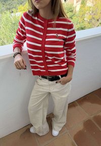 Vrouw draagt een rood-wit gestreepte gebuttonde trui, beige broek met wijde pijpen, zwarte riem en witte sneakers, staat op een betegelde vloer naast een witte reling.