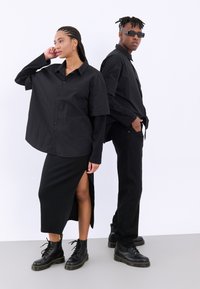 Camisa negra oversized de botones con mangas largas, falda negra midi con abertura lateral y botas negras con cordones. Diseño minimalista y monocrático.