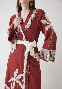Robe drapée bordeaux avec des détails en franges blanches et des motifs de feuilles. Elle est dotée d'une ceinture en tissu blanc nouée en nœud et de manches longues.