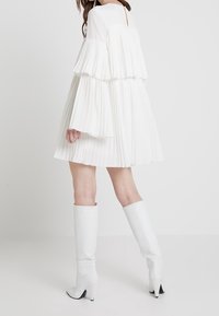 Robe blanche à volants et plissée avec des manches longues et un dos à ouverture en keyhole, associée à des bottes blanches à talons, arrivant jusqu'aux genoux.