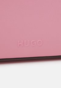 Texte en relief "HUGO" sur une surface en cuir rose doux, vue en gros plan avec un bord noir en bas.