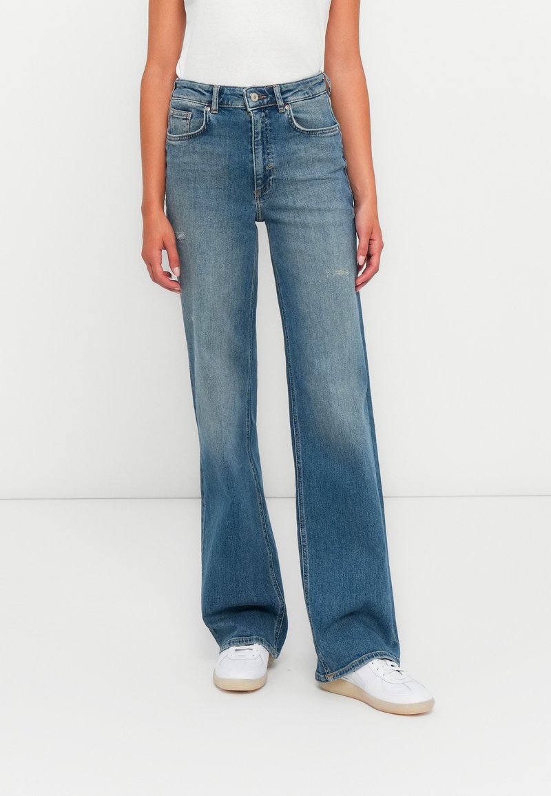 Pessoa a usar calças largas de cintura alta, azuis desbotadas, e sapatilhas brancas, de pé contra um fundo branco liso.