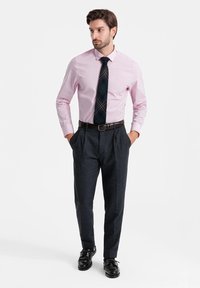 Camisa de manga larga rosa claro, corbata de cuadros oscuros, pantalones grises con pliegues, zapatos formales negros, de pie con las manos en los bolsillos frente a un fondo liso.